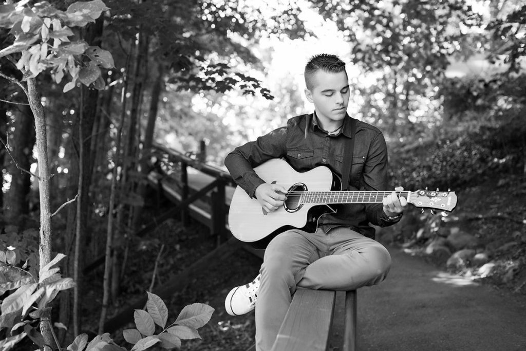 Black and white senior portrait of a teenage boy playing guitar on a wooded winding path, celebrating milestones.